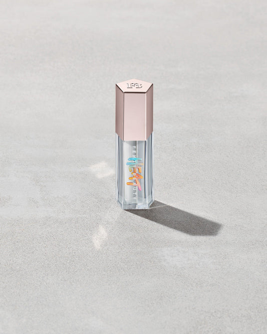 Closed tube of Gloss Bomb Heat lip gloss and plumper in the shade Glass Slipper on a grey background.