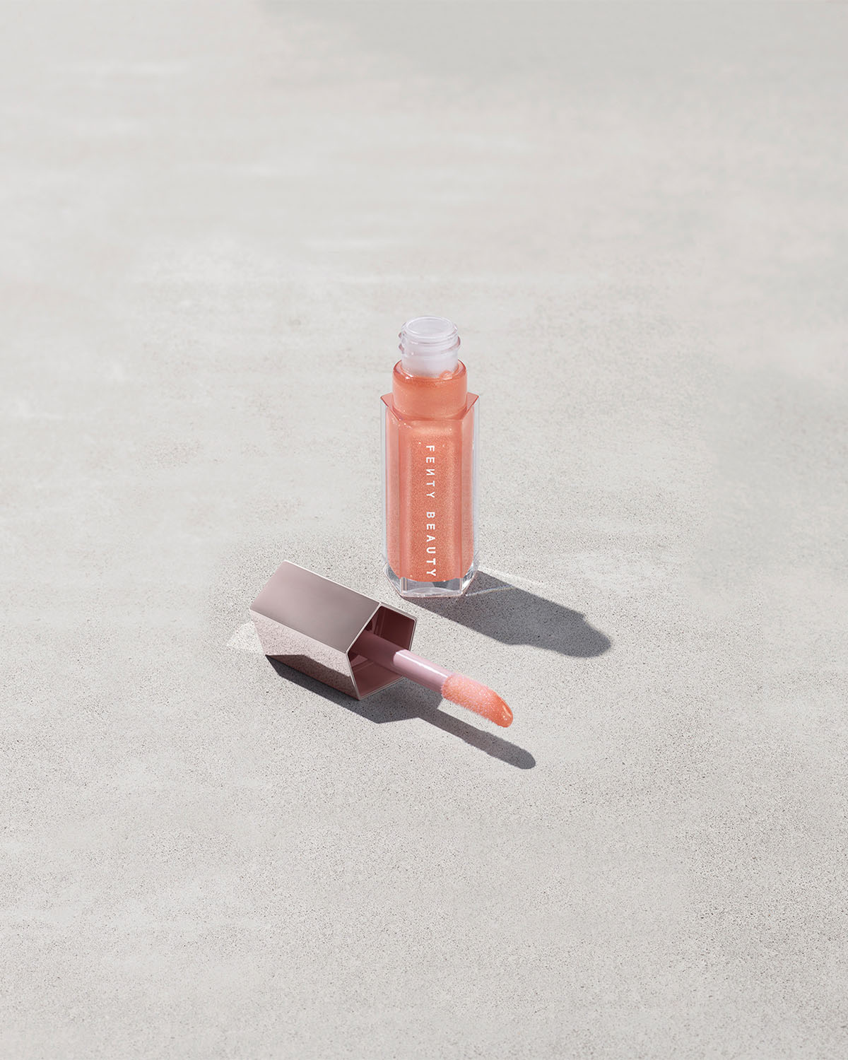 An open tube of Gloss Bomb Universal Lip Luminizer in the shade Champ Stamp Fantasy on a grey background.